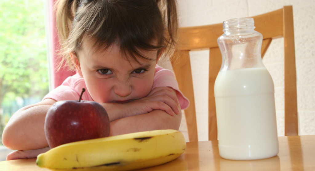Article on Picky Eating. Picture of toddler scowling in front of healthy foods, banana, apple and milk