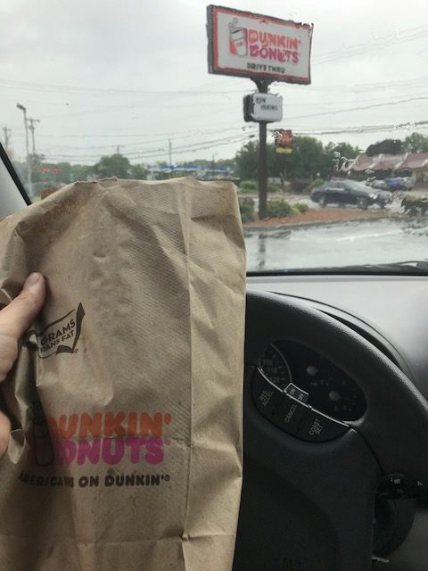 Dunkin' Donuts bag shot from the inside of a car with a Dunkin Donuts sign in the background. Author is on a Dunkin' Run.