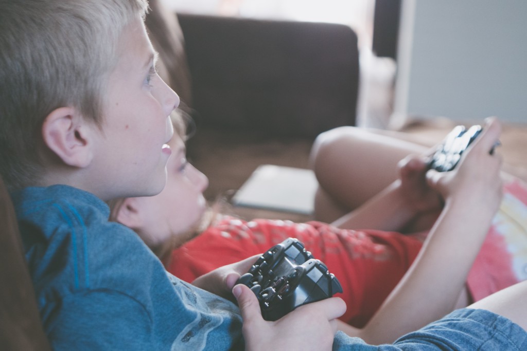 White boy with blond hair in foreground and white girl lounging in background. Both are holding video game controllers. Picture in Providence Moms Blog Article about playdate safety and questions parents should ask. 