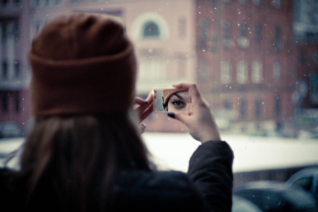Photo of woman looking at the reflection of her eye in a small mirror with a building and snow in the background. Providence Moms Blog post on self care.