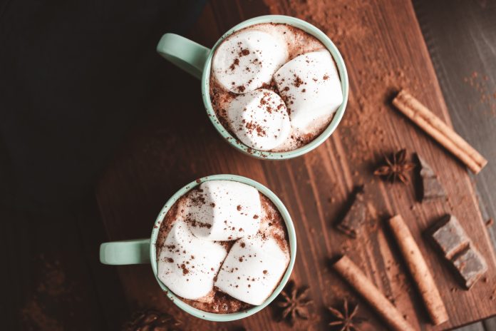 Hot chocolate drink with marshmallow in a cup on wooden board with cinnamon and star anise, top view.