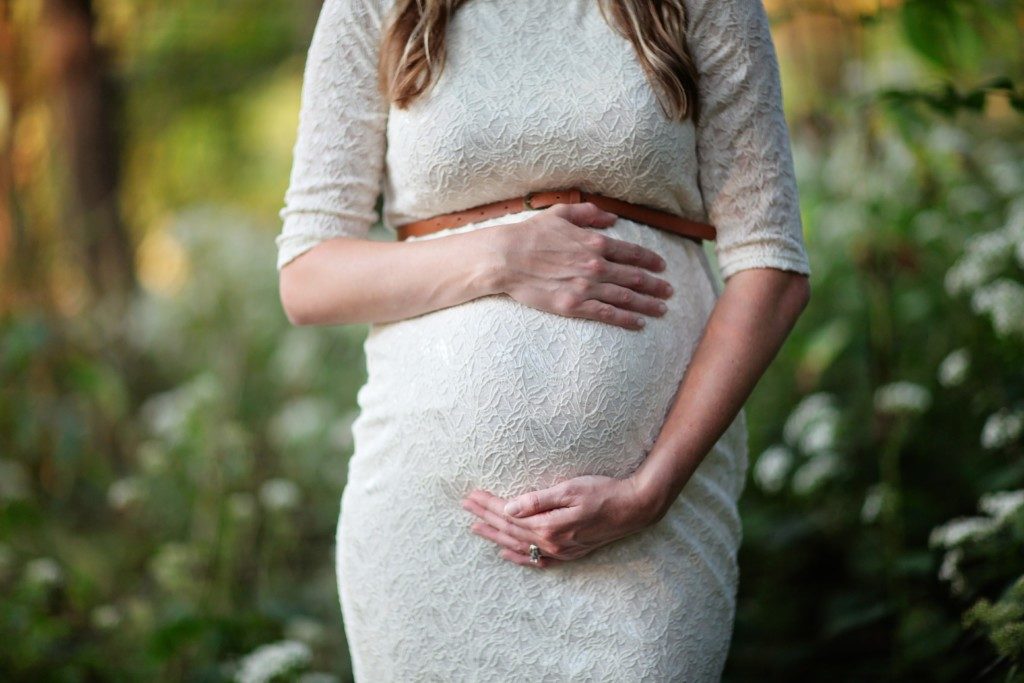 pregnant white woman wearing white lace dress against a background of greenery 