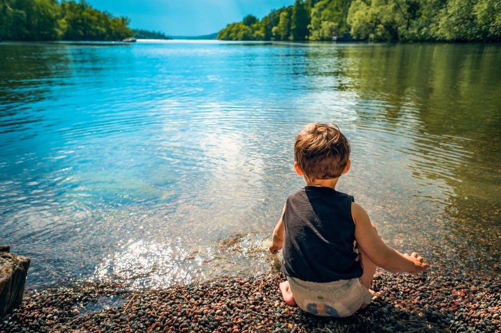 toddler sitting by a bright blue lake surrounded by trees