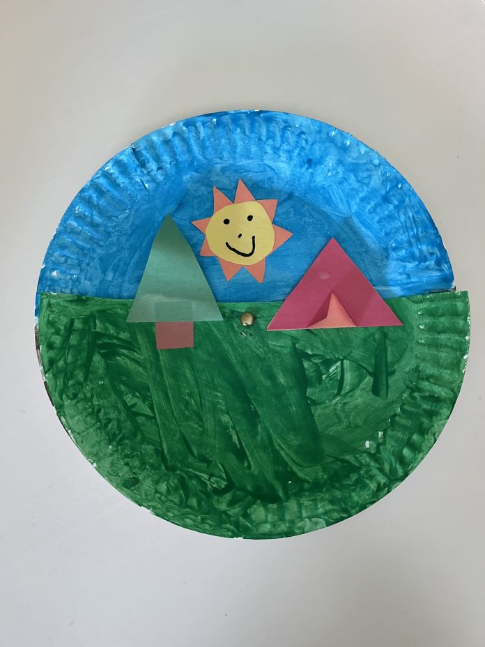 A paper plate with a half a paper plate attached together with a split pin with a tree, tent and sun made out of a paper stuck on to make a camping scene