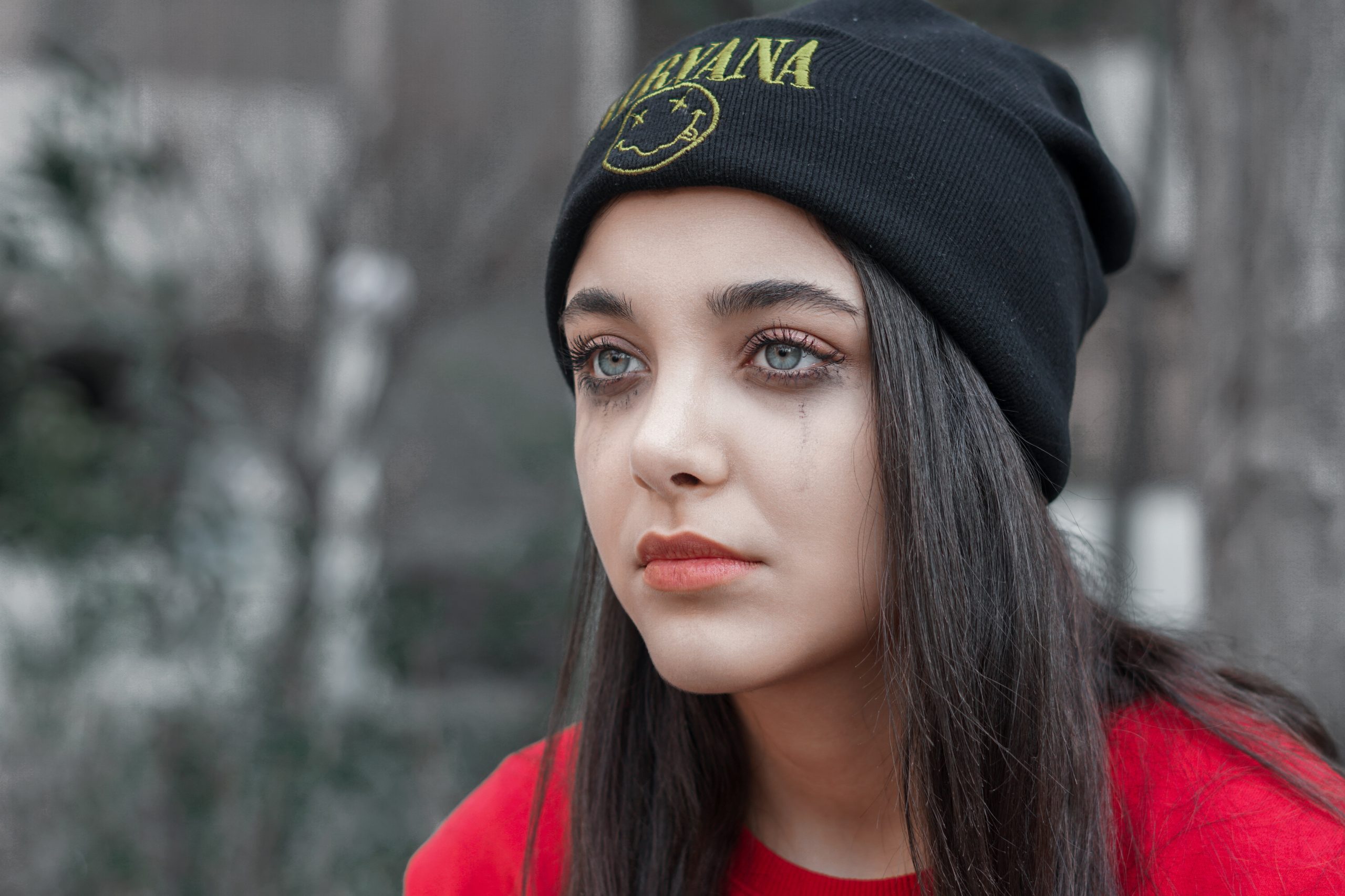 A white woman with pale skin and long, straight brunette hair stares out into space looking melancholy. She is wearing a black "Nirvana" beanie hat and has dark eye make up that is running down her face leaving dirty looking trails on her cheeks