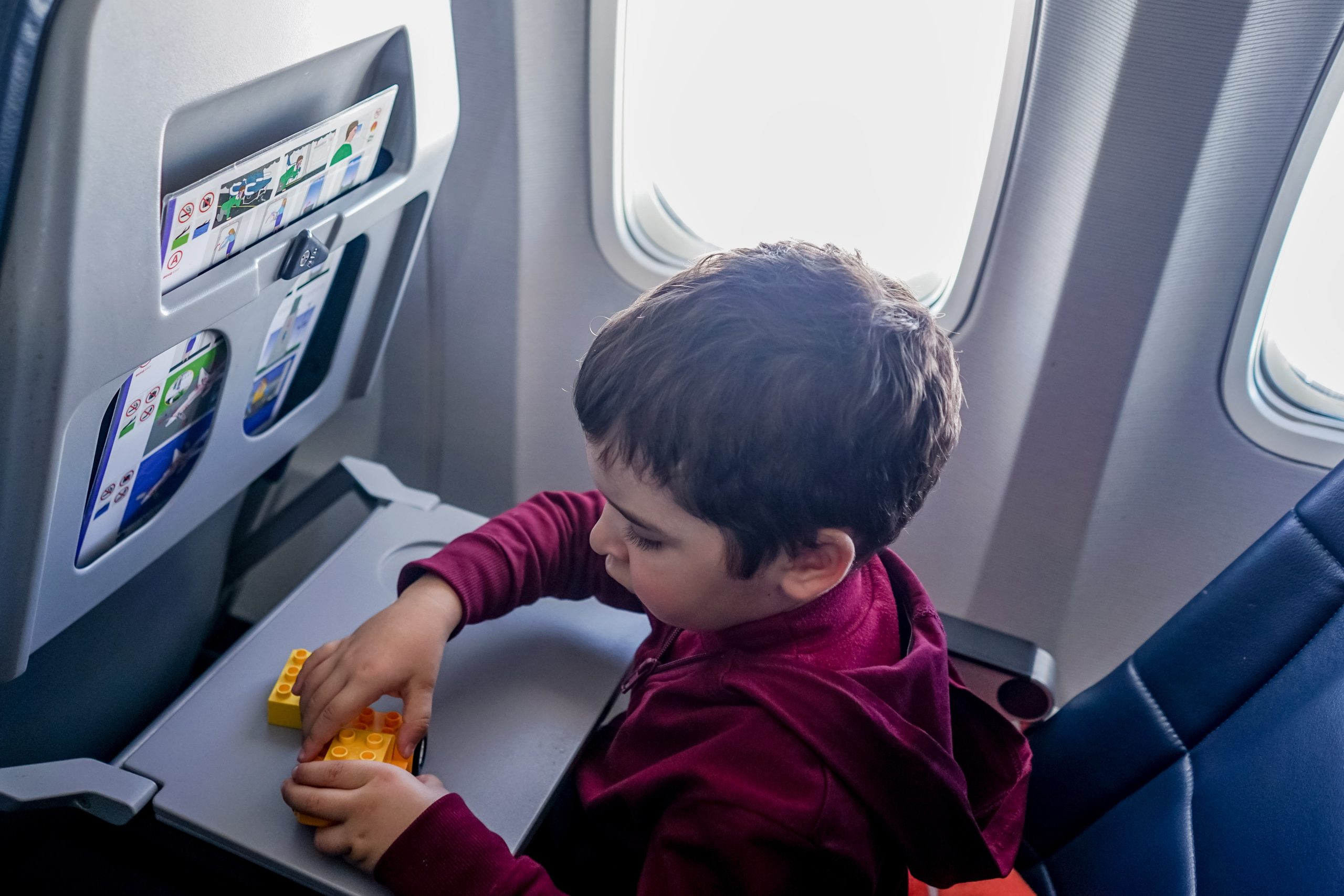 Flying with toddlers: A little boy sits in airplane window seat with the tray table down. he is playing with duplo blocks 