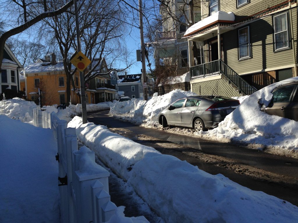 A snowy street with a car parked between piles of snow that are higher than the car
