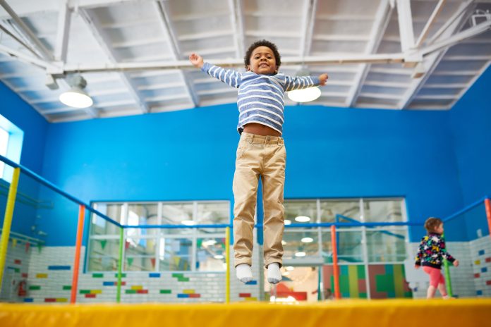 Full length low angle portrait of happy African-American boy jumping on trampoline in colorful kids play center, copy space